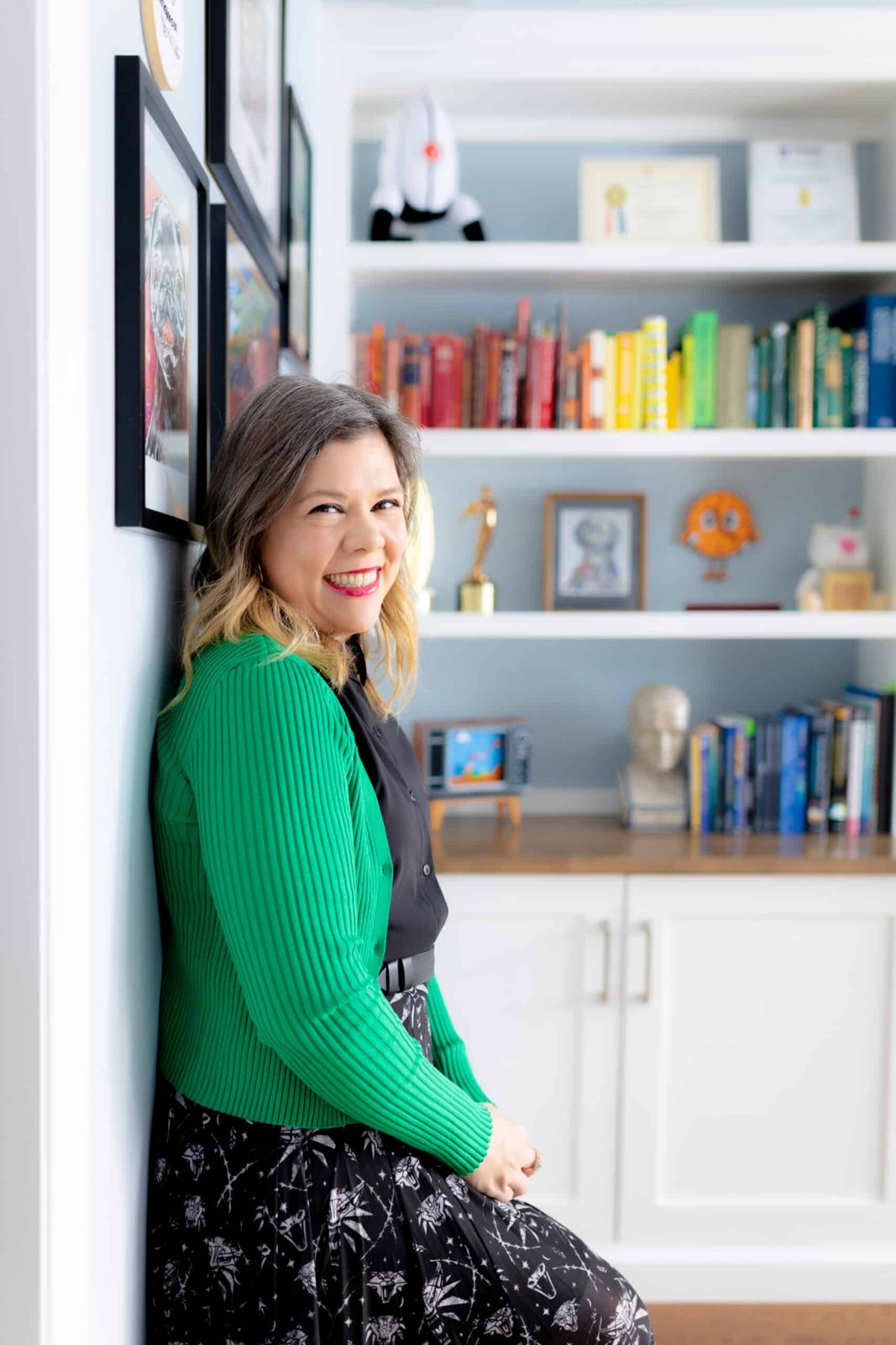 Rachel Kowert smiling in a green cardigan, leaning against a wall beside a bookshelf full of books and game figures