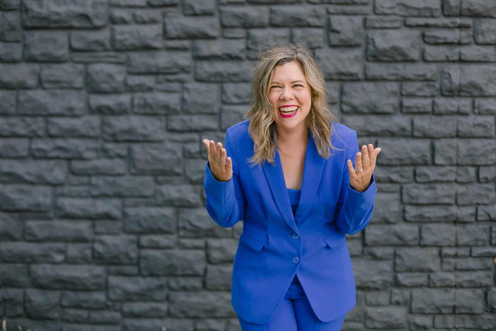 Rachel Kowert laughing and gesturing in a bright blue suit in front of a dark stone wall