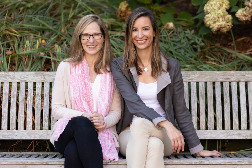Carrie James and Emily Weinstein sitting together on a wooden park bench, smiling