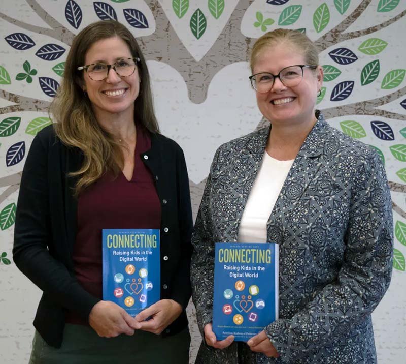 Jenny Radesky and Megan Moreno standing together in front of a mural, each holding a copy of Connecting