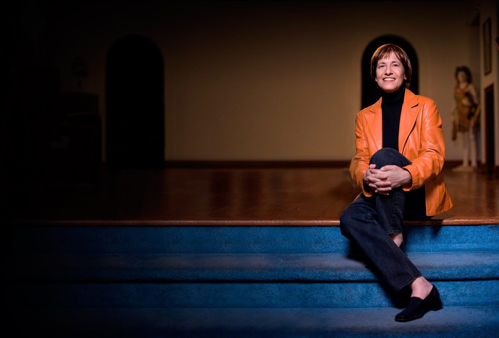 Kathy Hirsh-Pasek in an orange leather jacket, sitting on the steps of a dark stage