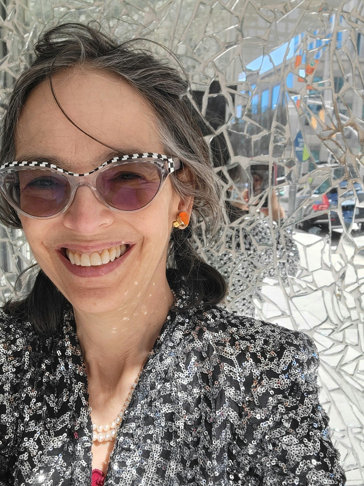 Lenore Skenazy, smiling in sunglasses with white frames and a sequined jacket, in front of a mirrored wall