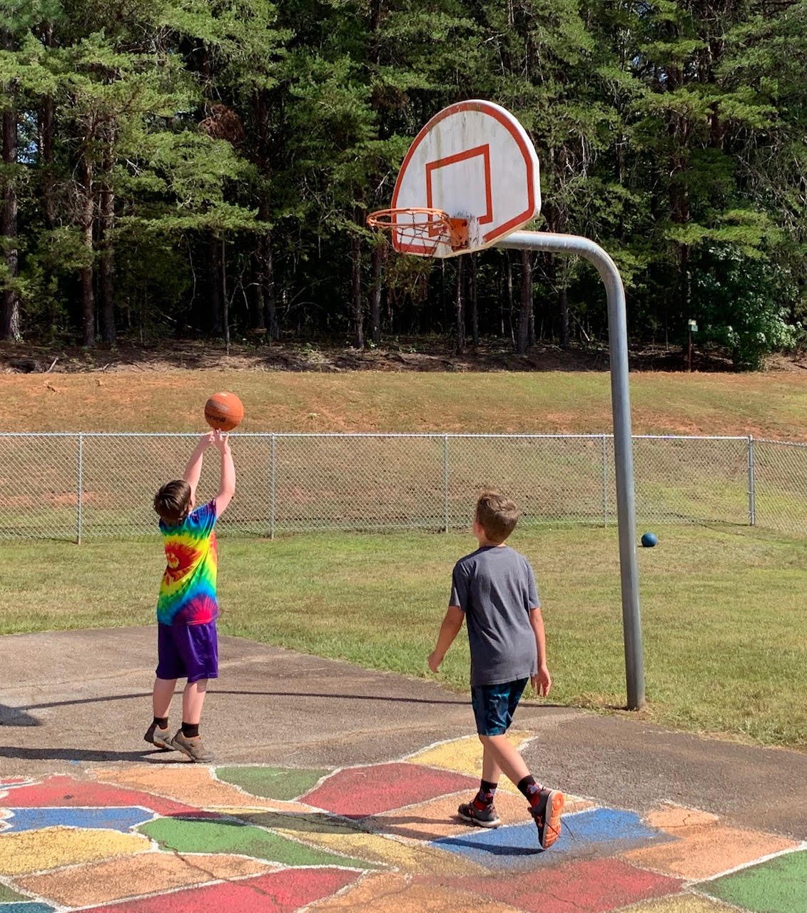 Two boys shooting baskets on an outdoor court with colorful painted squares, trees behind them