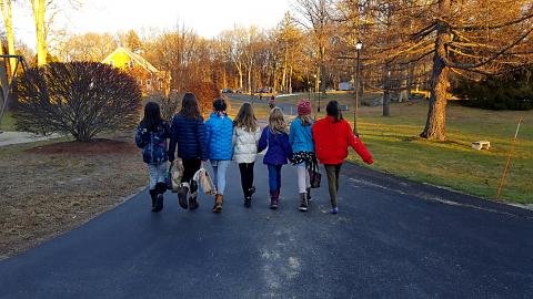 A group of children and teens in winter coats walking together up a paved drive under bare trees