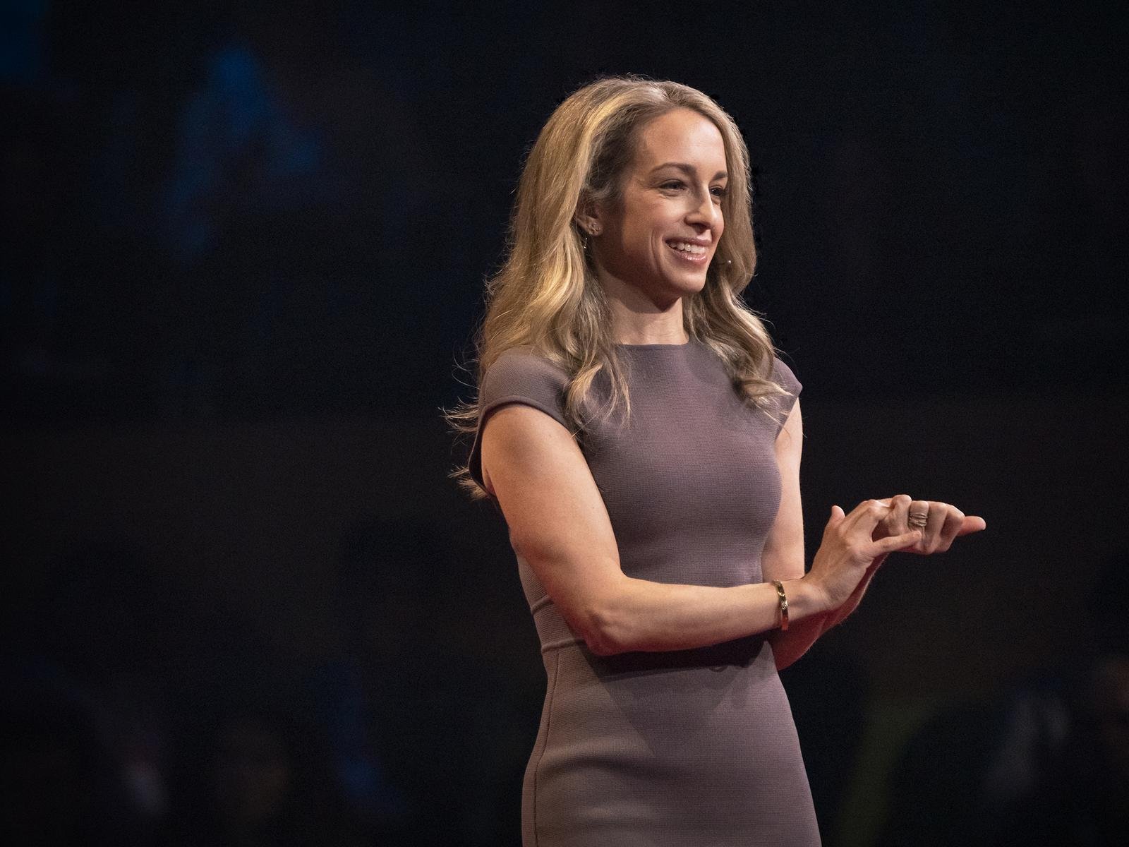 Becky Kennedy smiling on the TED stage in a gray dress, hands together as she speaks
