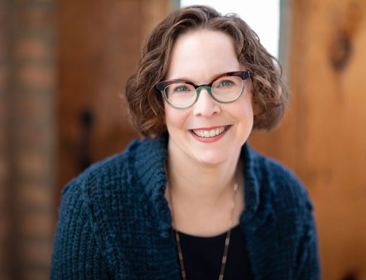 Devorah Heitner smiling in glasses and a dark knit cardigan, wood paneling behind her