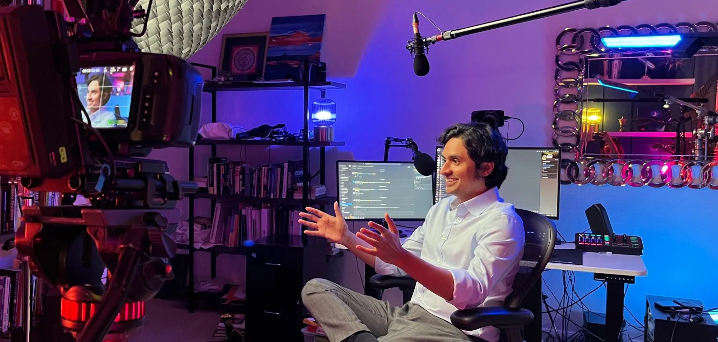 Alok Kanojia gesturing as he talks in front of a camera in his home streaming studio, lit in purple and blue