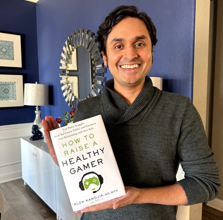 Alok Kanojia smiling and holding up a copy of How to Raise a Healthy Gamer in his living room