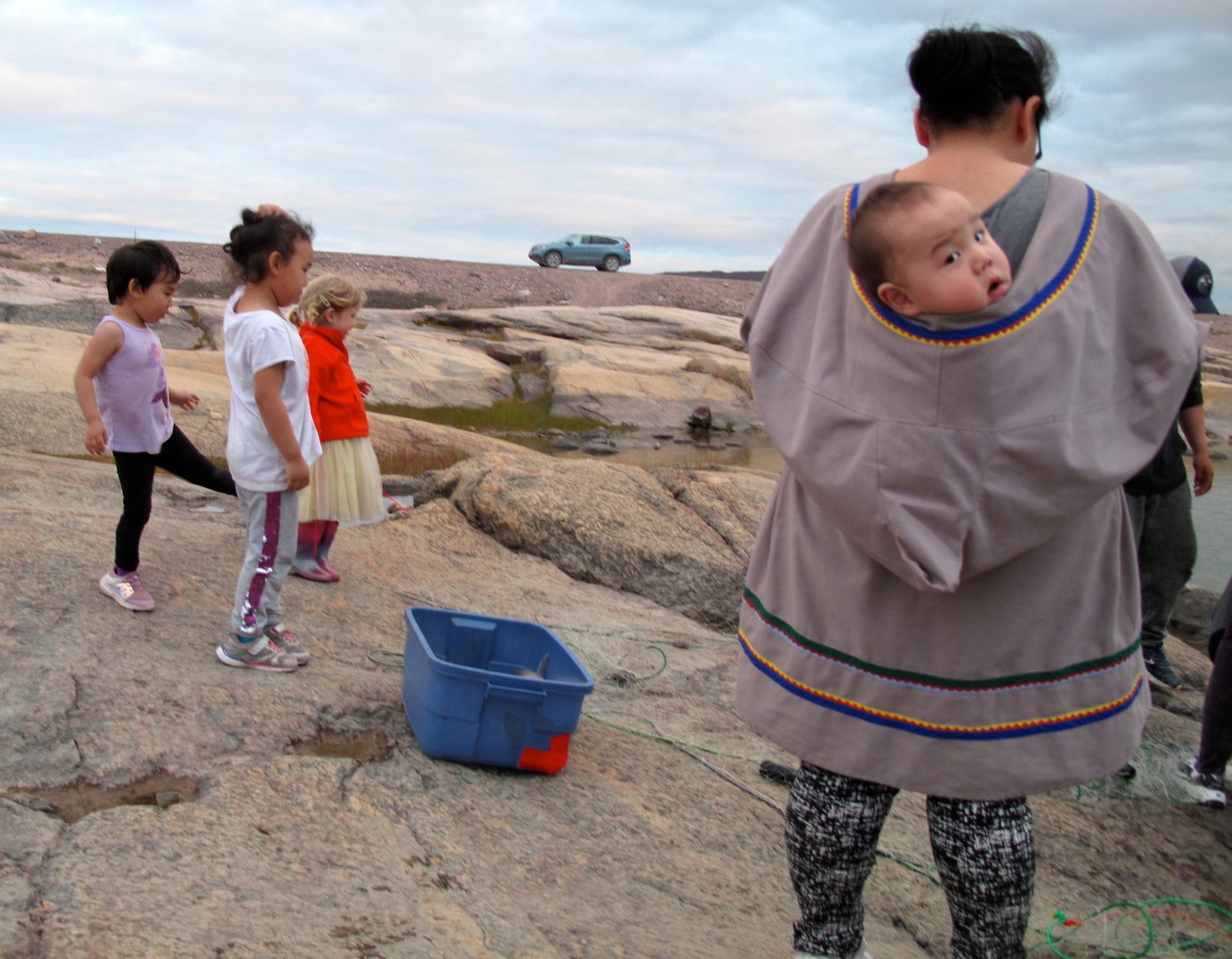 Three small children playing on flat Arctic rocks by the water while a woman stands nearby with a baby peeking out of the hood of her parka