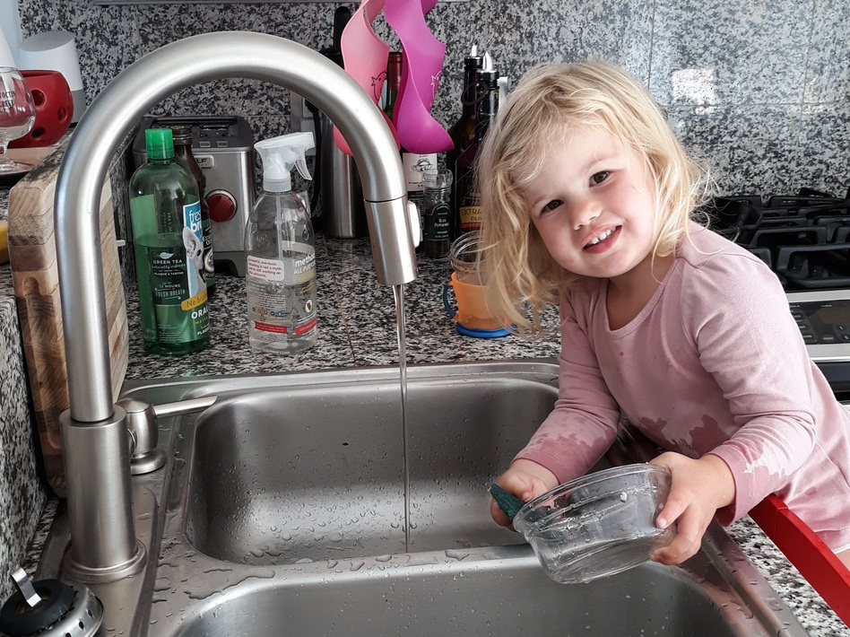 A blonde toddler in pajamas smiling while she rinses a glass bowl under the kitchen faucet
