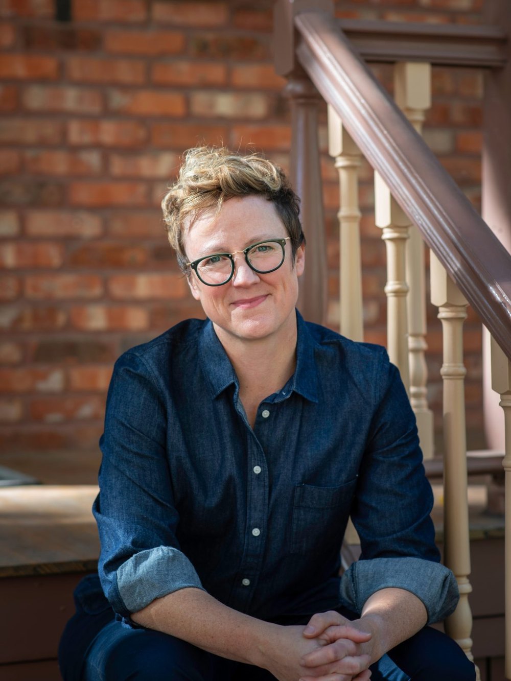 Erin Walsh, with short blond hair and glasses, sitting on a stair and smiling slightly, in a denim shirt