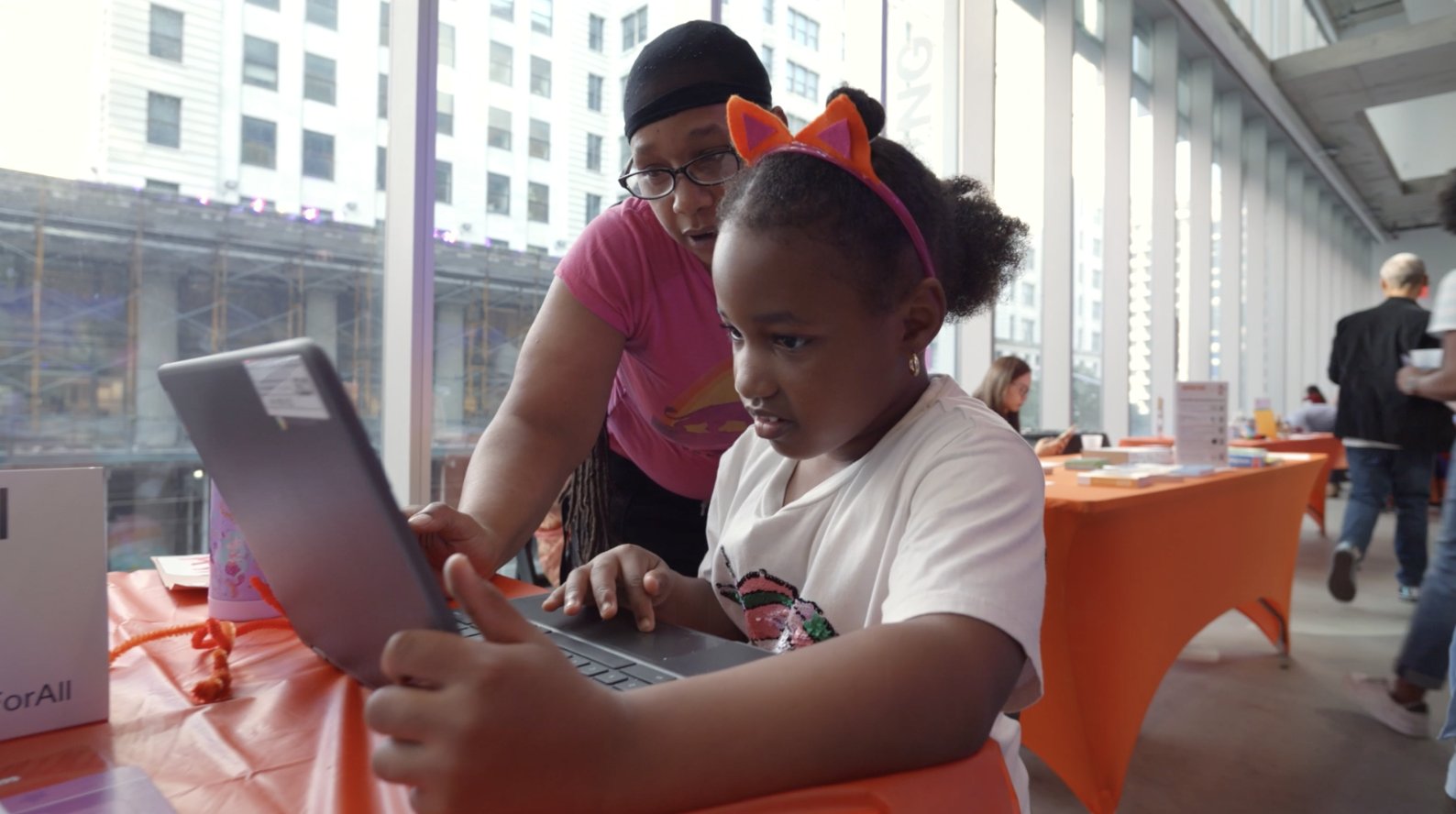 A girl wearing cat ears types on a laptop at a table while a woman leans over to help her