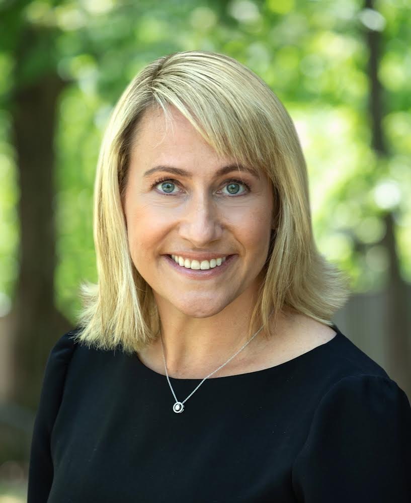Phyllis Fagell smiling outdoors, with shoulder-length blond hair and a black top, trees behind her