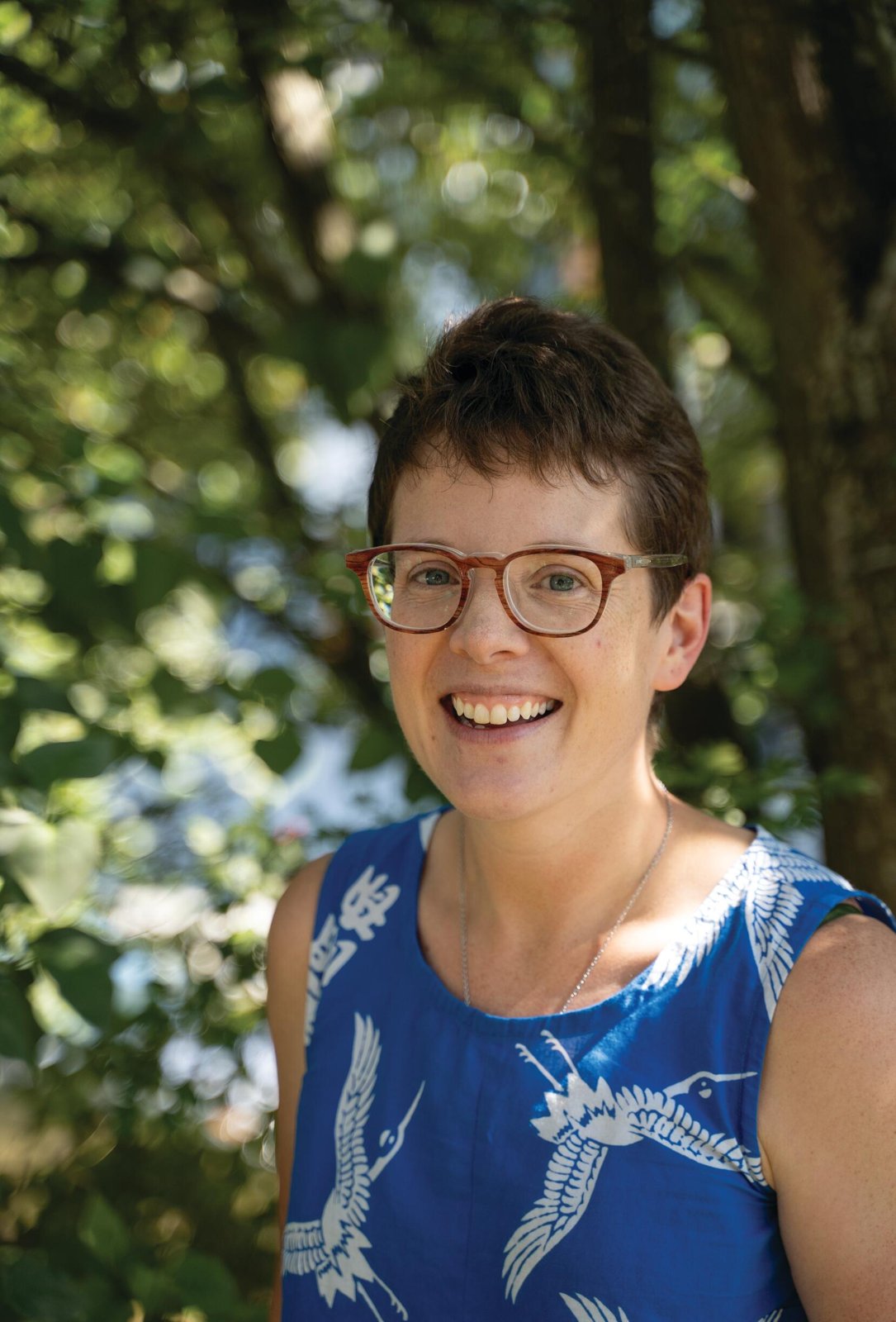 Ash Brandin smiling in glasses and a blue sleeveless top printed with cranes, in front of green trees