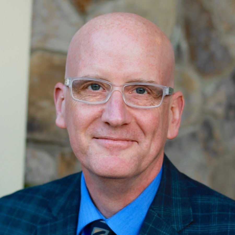 Daniel Willingham smiling in a blue shirt, tie and plaid jacket in front of a stone wall