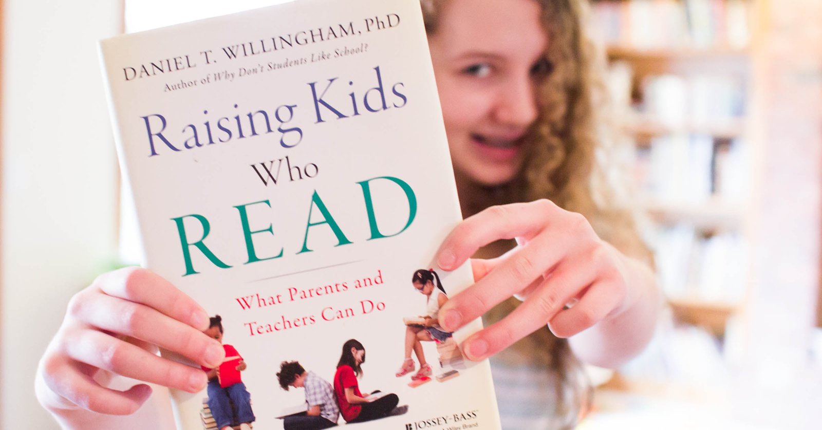 A girl with curly hair holding up a copy of Raising Kids Who Read in front of a bookshelf