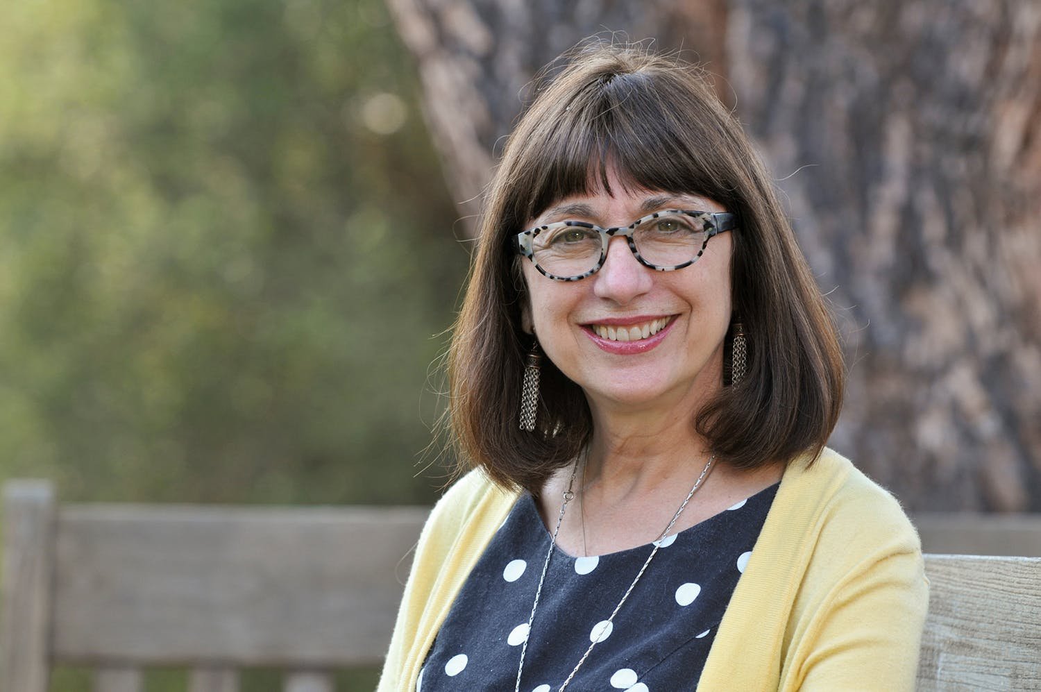 Maryanne Wolf in glasses and a yellow cardigan, smiling on a bench under a large tree