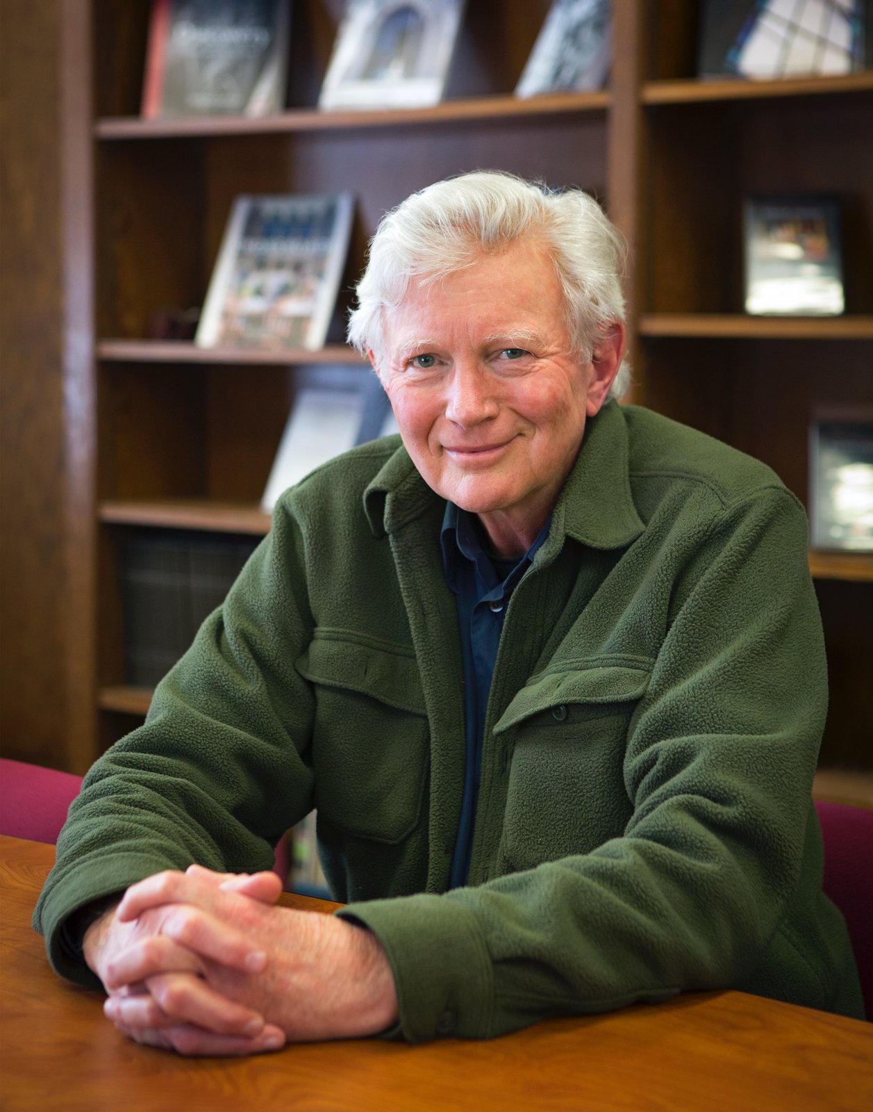 Peter Gray, white-haired and smiling, sitting at a table with his hands folded in front of a bookshelf