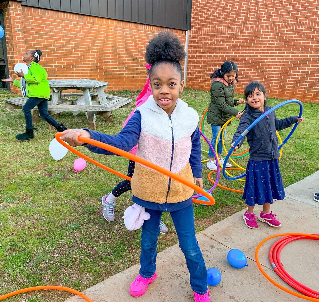 A girl in a pink and white jacket spinning an orange hula hoop on the grass outside a school, other kids playing with hoops and balloons behind her