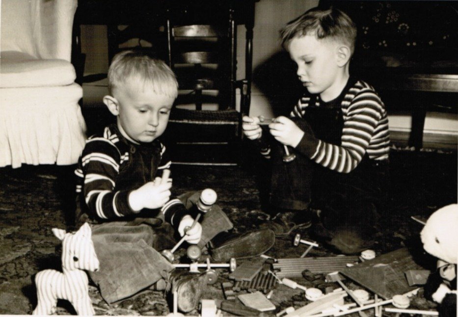 A black-and-white photo of two small boys in striped sweaters building with Tinker Toys on a living-room rug