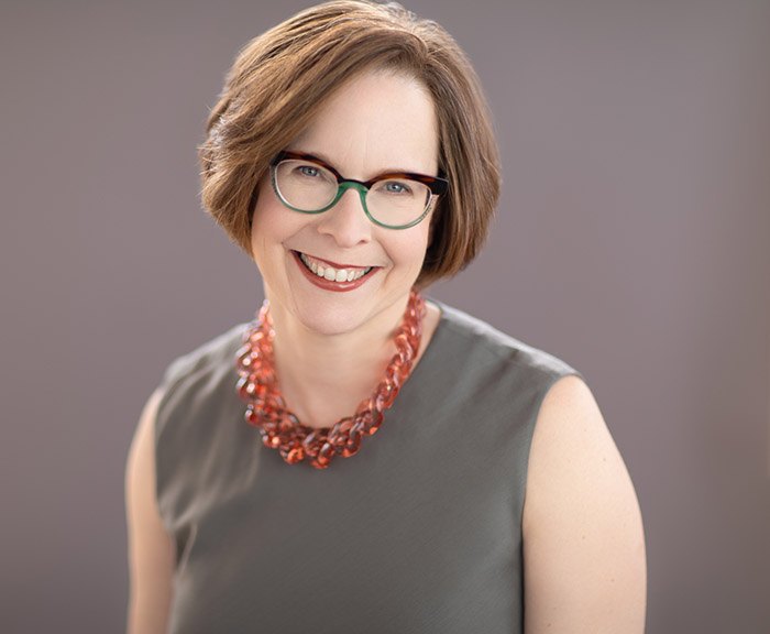 Devorah Heitner smiling, in glasses and a coral necklace, against a gray backdrop
