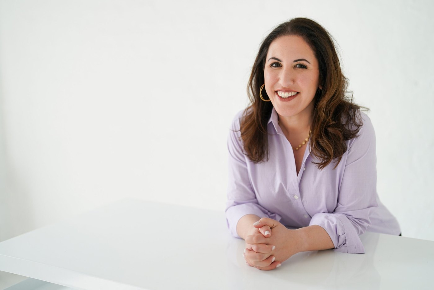 Ana Homayoun smiling, seated at a white table with her hands folded, in a lavender blouse