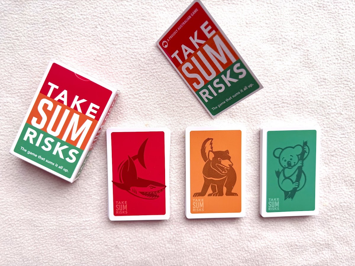 The Take Sum Risks box and rules card above three face-down piles: a red pile with a shark, an orange pile with a Tasmanian devil and a green pile with a koala