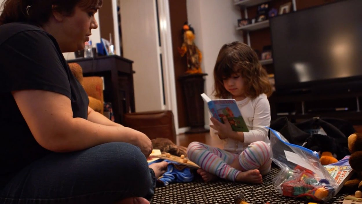 A young girl sitting on the floor reading a picture book while her mother sits beside her