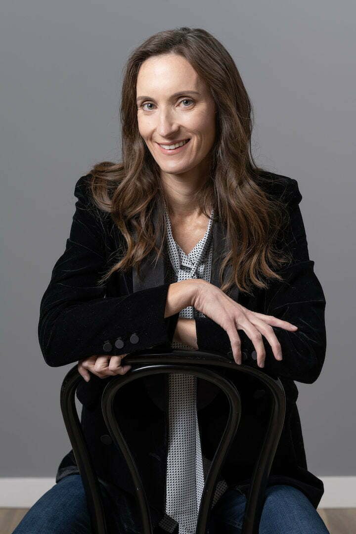 Katie Davis sitting backward on a wooden chair in a dark velvet jacket, smiling, against a gray studio backdrop