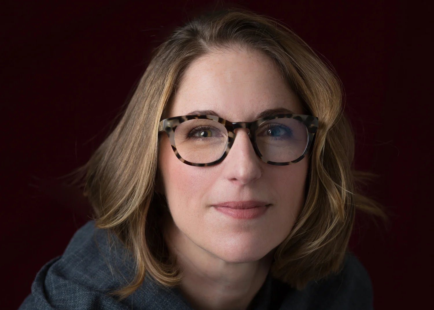 Jessica Lahey, in tortoiseshell glasses and a gray top, against a dark red background