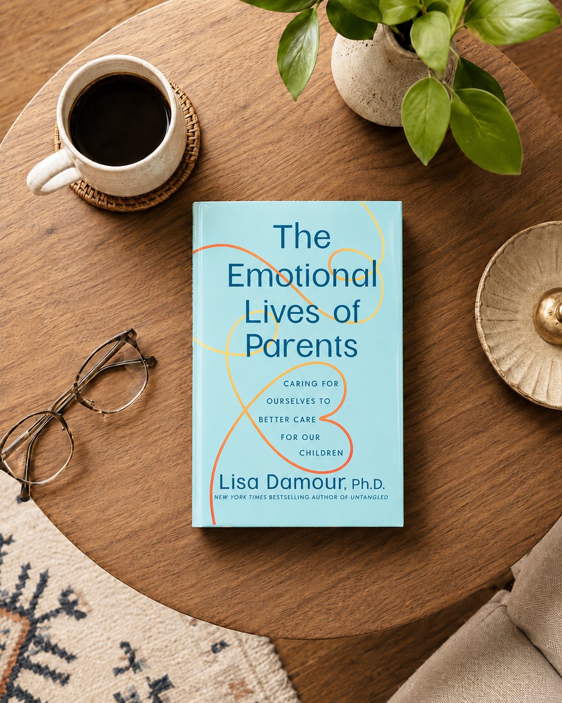 The Emotional Lives of Parents by Lisa Damour, a pale blue hardcover with orange and yellow looping lines, on a wooden table beside a coffee mug, glasses and a plant
