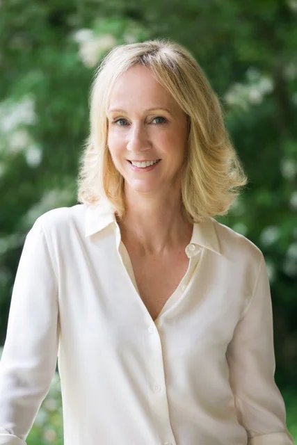 Meghan Cox Gurdon smiling in a cream blouse, with green trees behind her