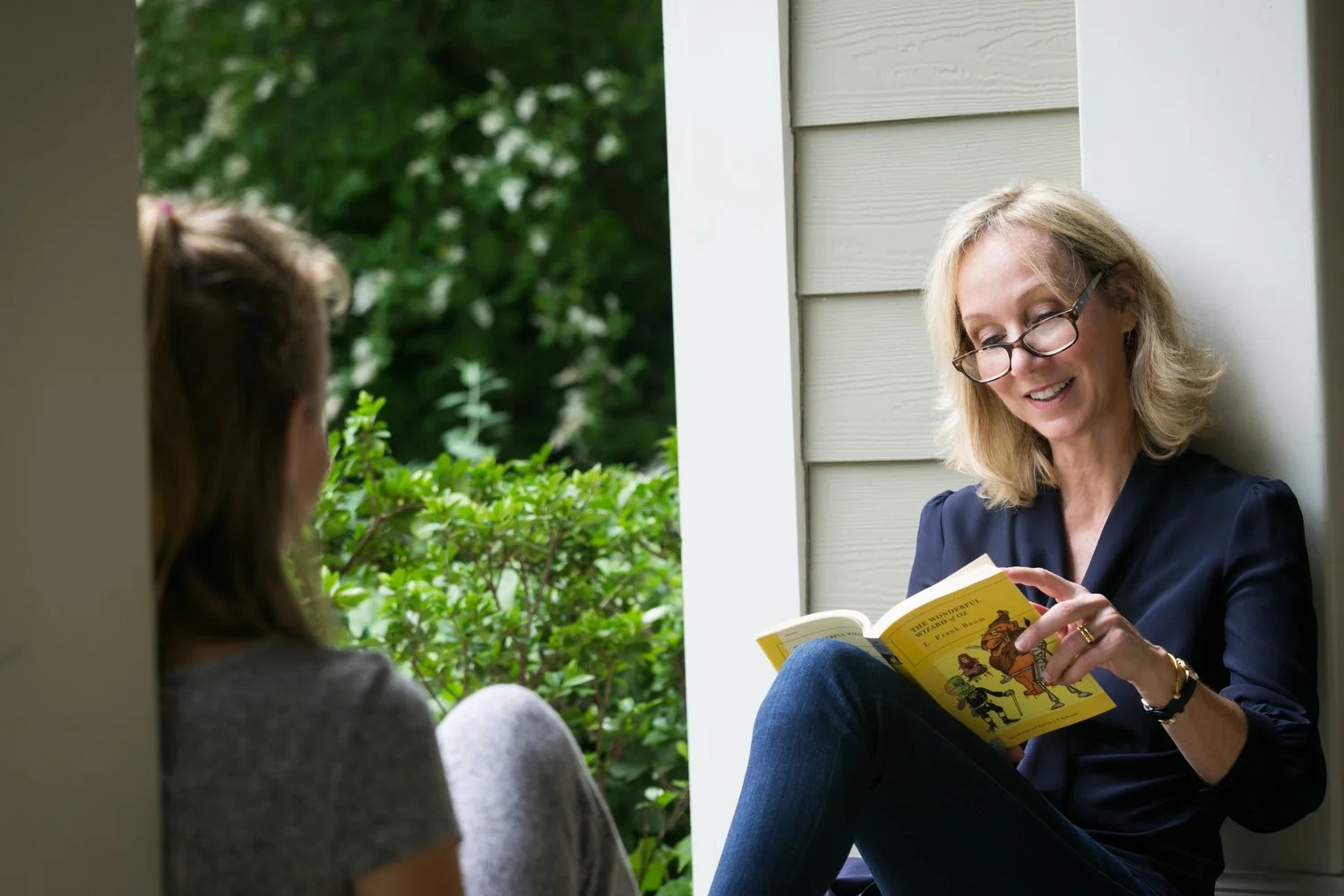Meghan Cox Gurdon sitting on a porch step reading The Wonderful Wizard of Oz aloud to a girl sitting across from her