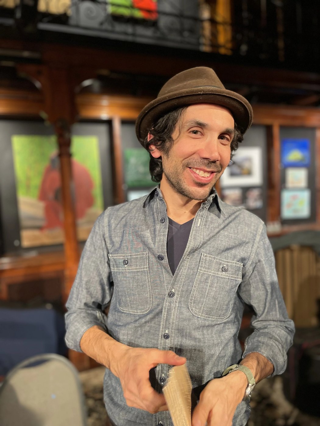 Jordan Shapiro smiling in a brown fedora and a gray button-up shirt, in a wood-paneled room