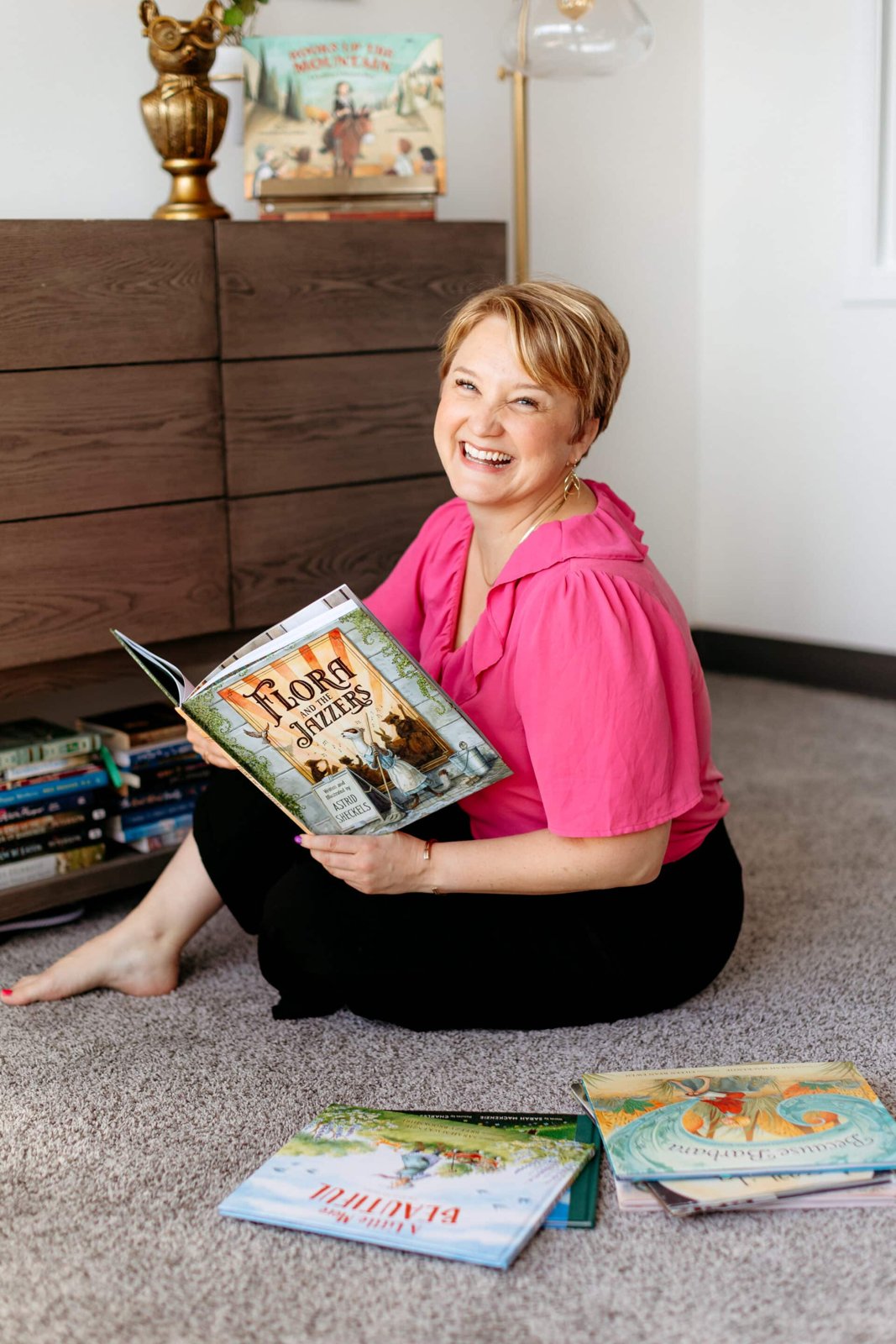 Sarah Mackenzie laughing on the floor with a picture book open in her lap and more picture books spread around her, including her own A Little More Beautiful