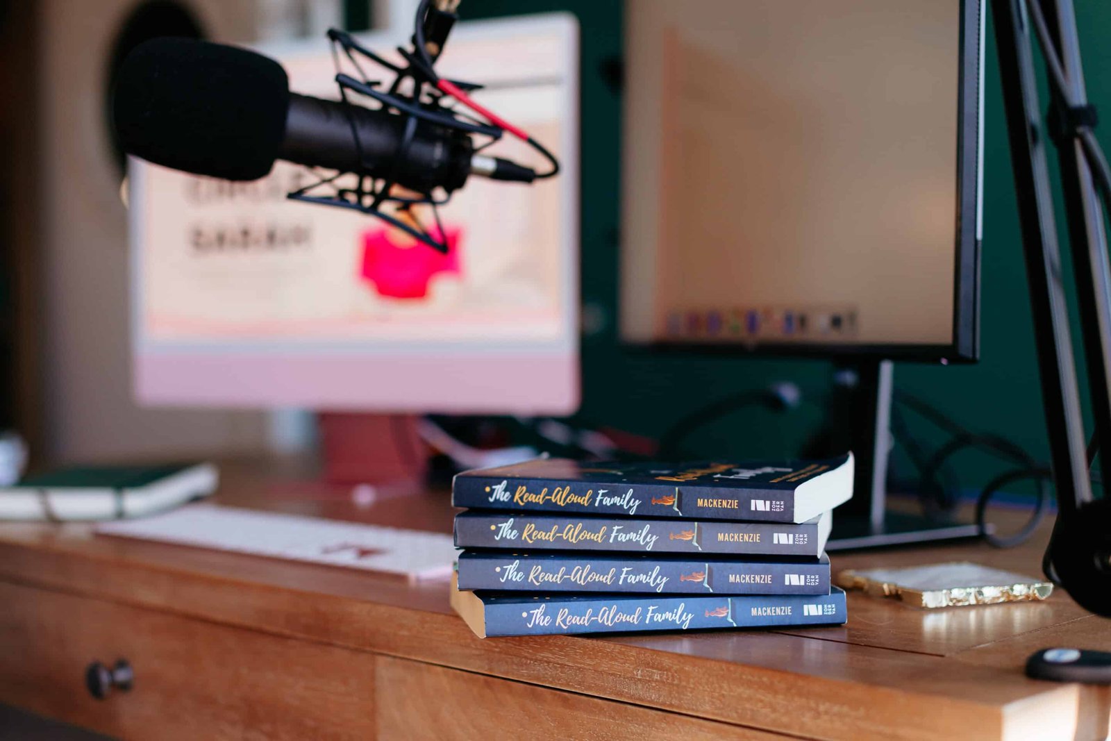 A stack of copies of The Read-Aloud Family on a desk beside a podcast microphone and a computer screen