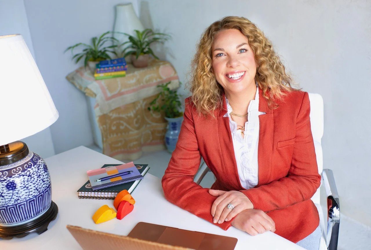 Tina Payne Bryson, smiling in a red blazer, sitting at a white desk with colored pencils and a lamp