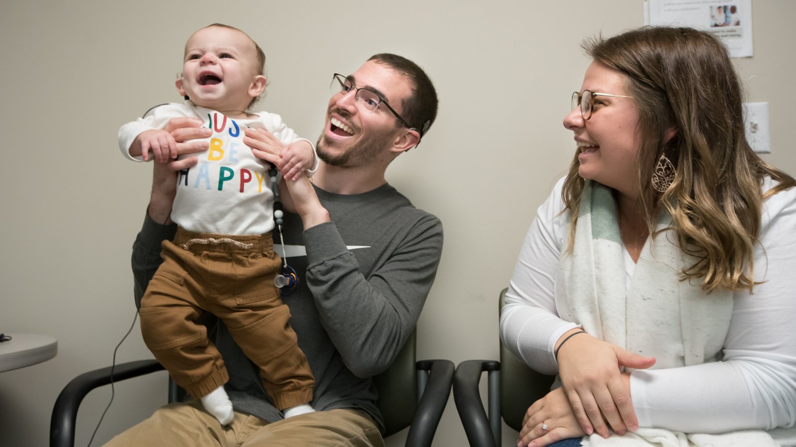 A smiling baby with a cochlear implant, held up by his laughing father as his mother looks on in a clinic room