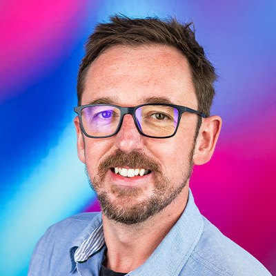 Pete Etchells smiling in glasses and a blue shirt against a pink and blue background