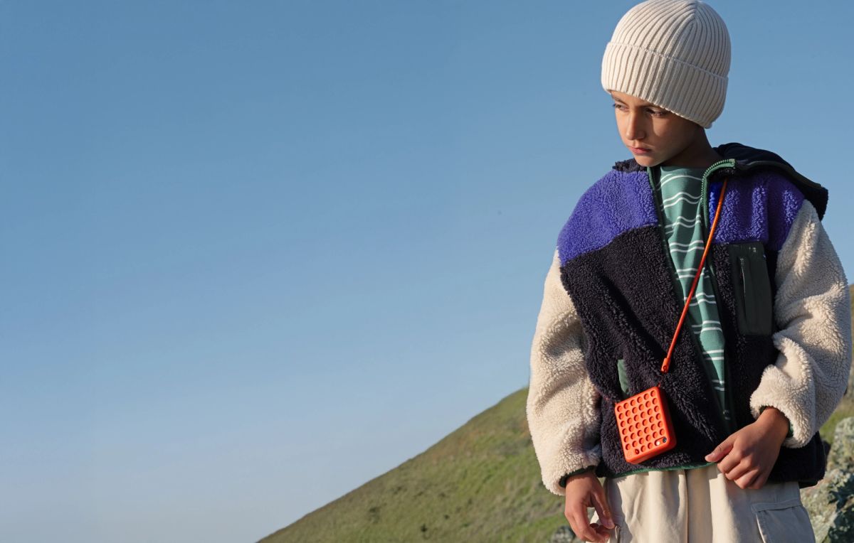 A boy on a grassy hillside wearing an orange Freckle on a cross-body strap over a fleece jacket
