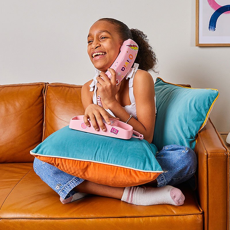 A laughing girl on a tan leather couch holding a pink handset covered in stickers, the phone's base on a teal pillow in her lap