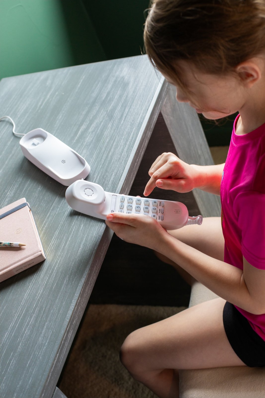 A girl in a pink shirt dialing the keypad on the inside of a white trimline handset, its base on a gray desk
