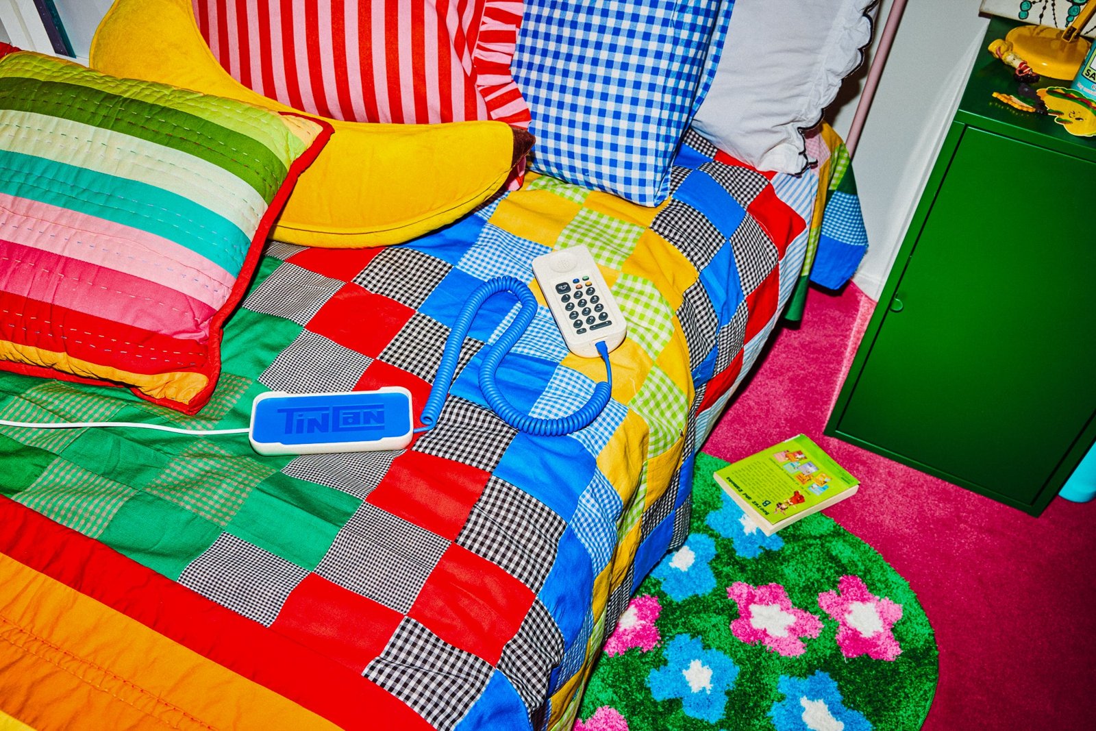 A white Tin Can lying on a patchwork quilt, keypad facing up, its blue curly cord running to a small blue Tin Can base