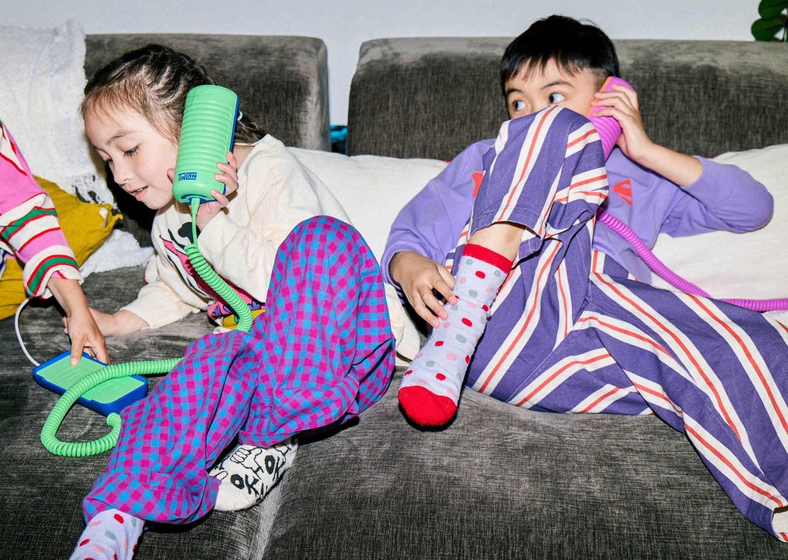 Two kids sprawled on a gray couch in pajamas, each holding a Tin Can to their ear, one green and one pink, with the curly cords running to their bases