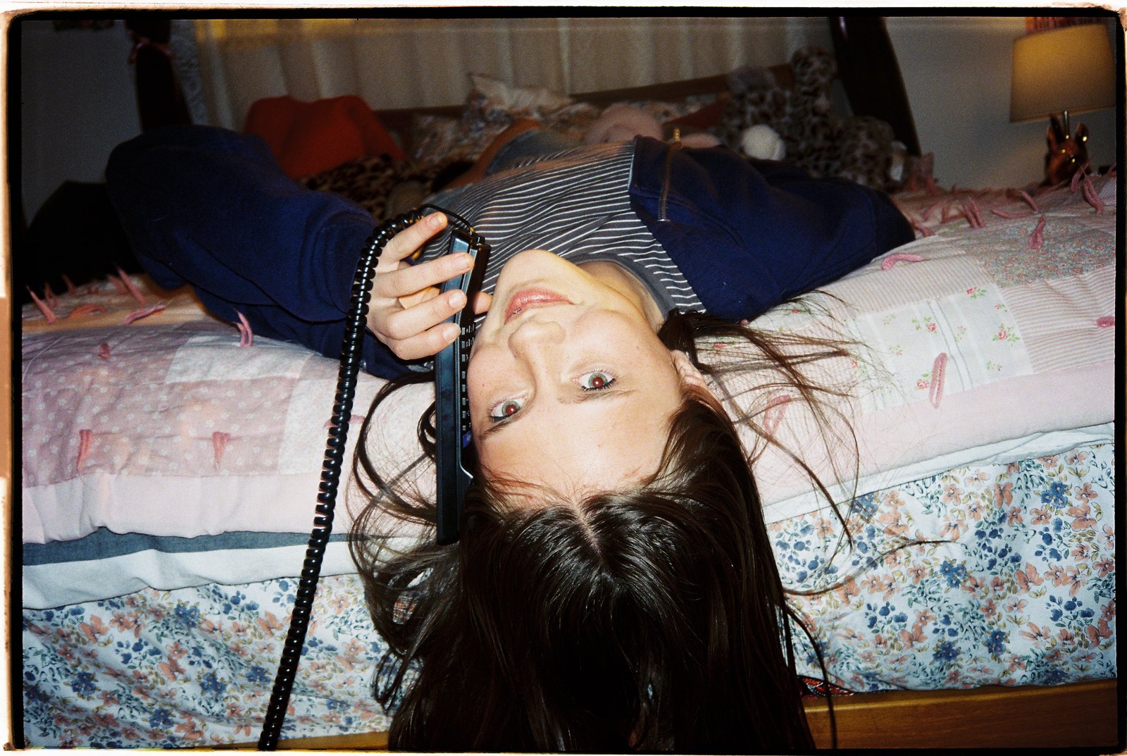 A girl lying upside down across a bed with a floral quilt, holding a black corded handset to her ear