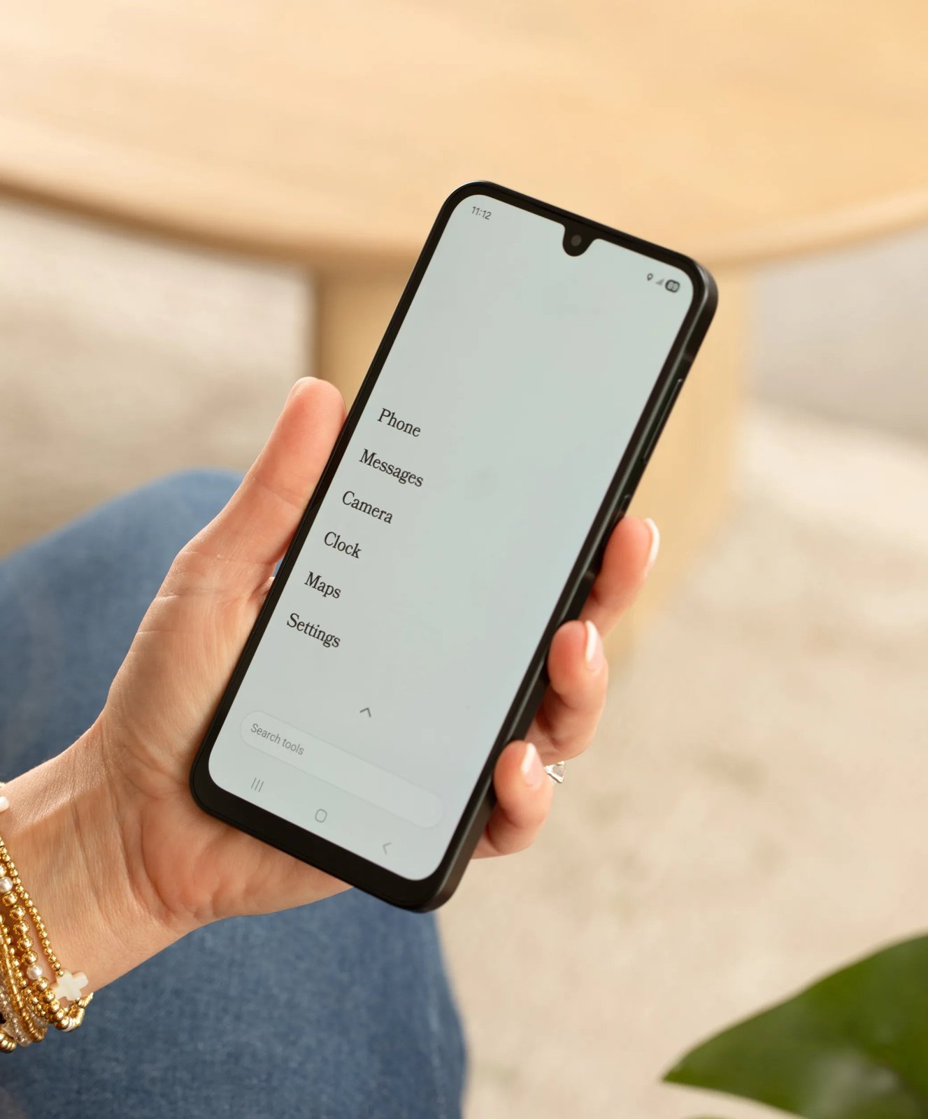 A hand holding a black Wisephone II over a pair of jeans, the screen showing the WiseOS list of Phone, Messages, Camera, Clock, Maps and Settings
